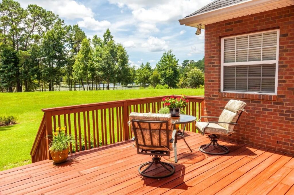 Timber deck with railing attached to a brick home, overlooking a green lawn and trees